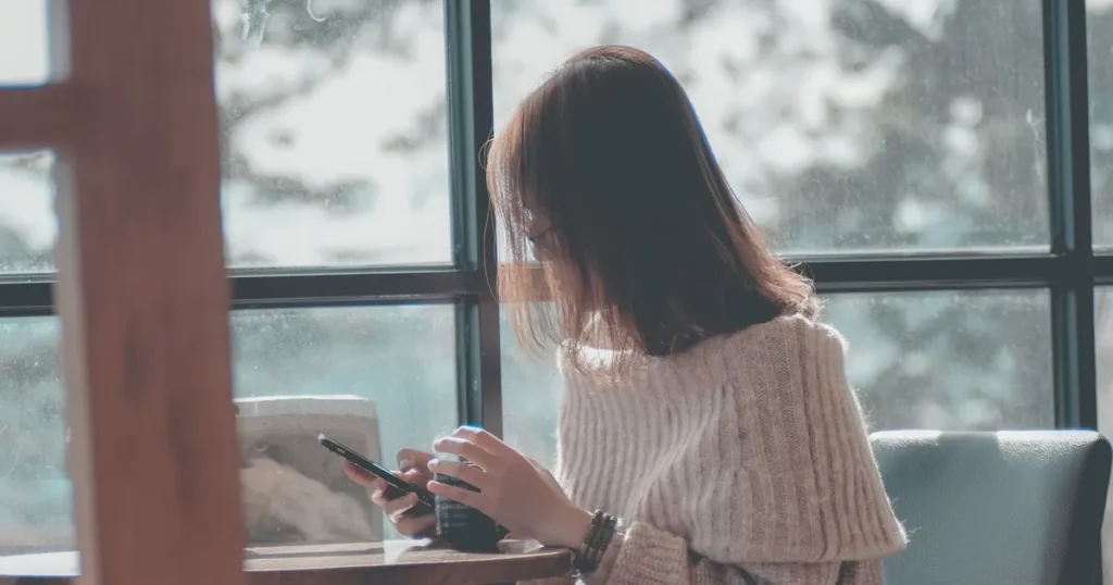 Woman at a cafe window checking her phone for the realistic ways to get money now in Canada